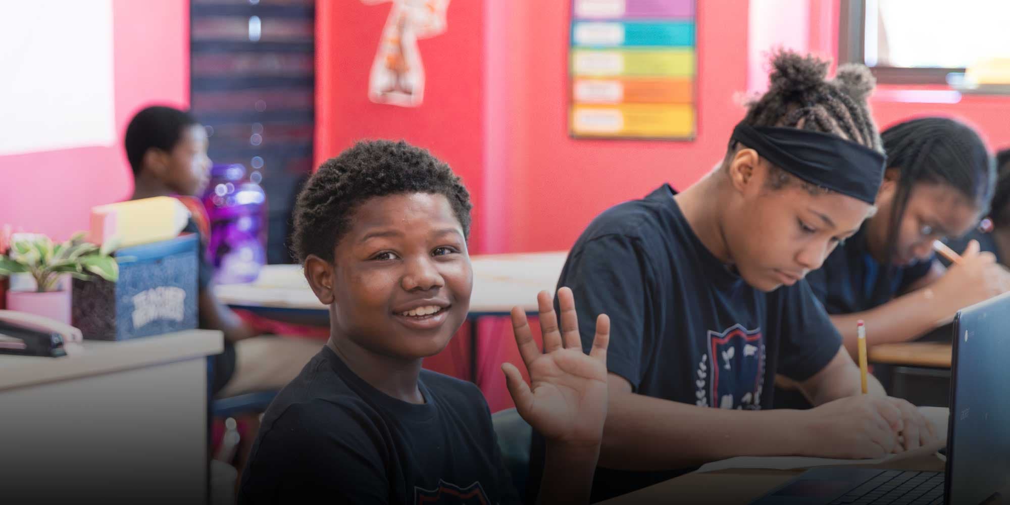 Smiling student waving from a desk in a classroom while classmates work.