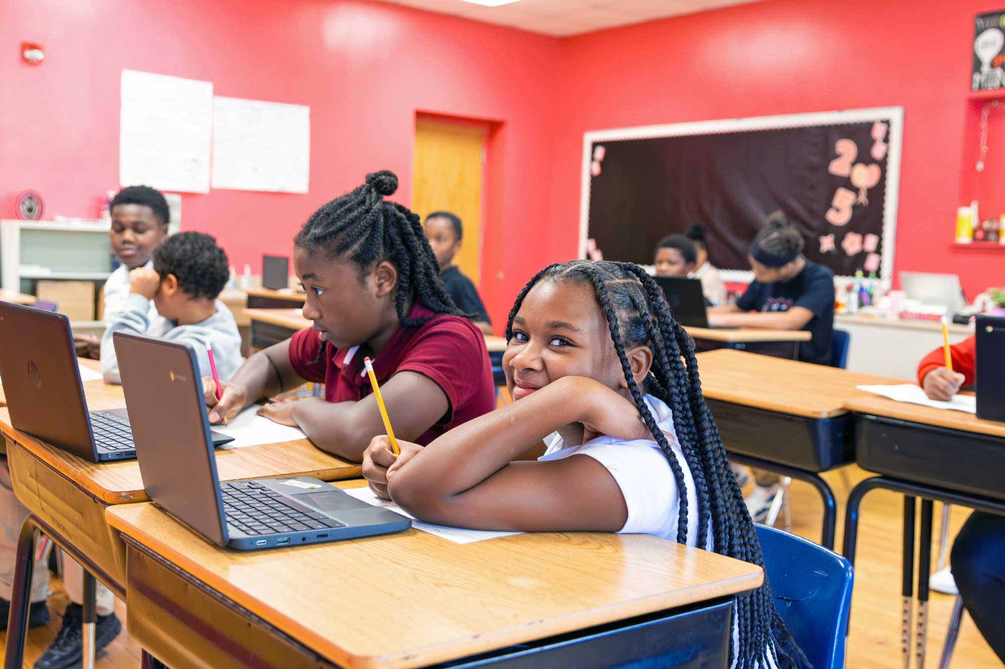 Students writing and working on laptops at desks in a classroom with red walls.
