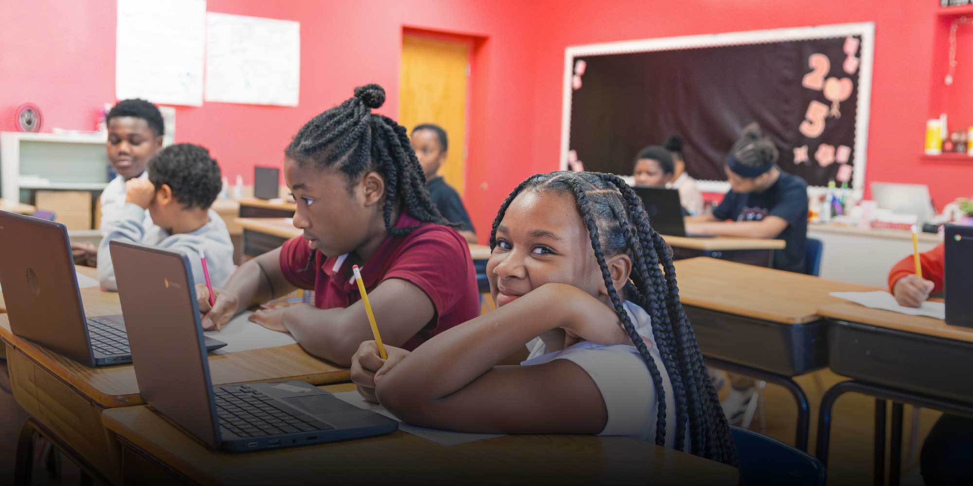 Students working on laptops at classroom desks, one smiling at the camera.