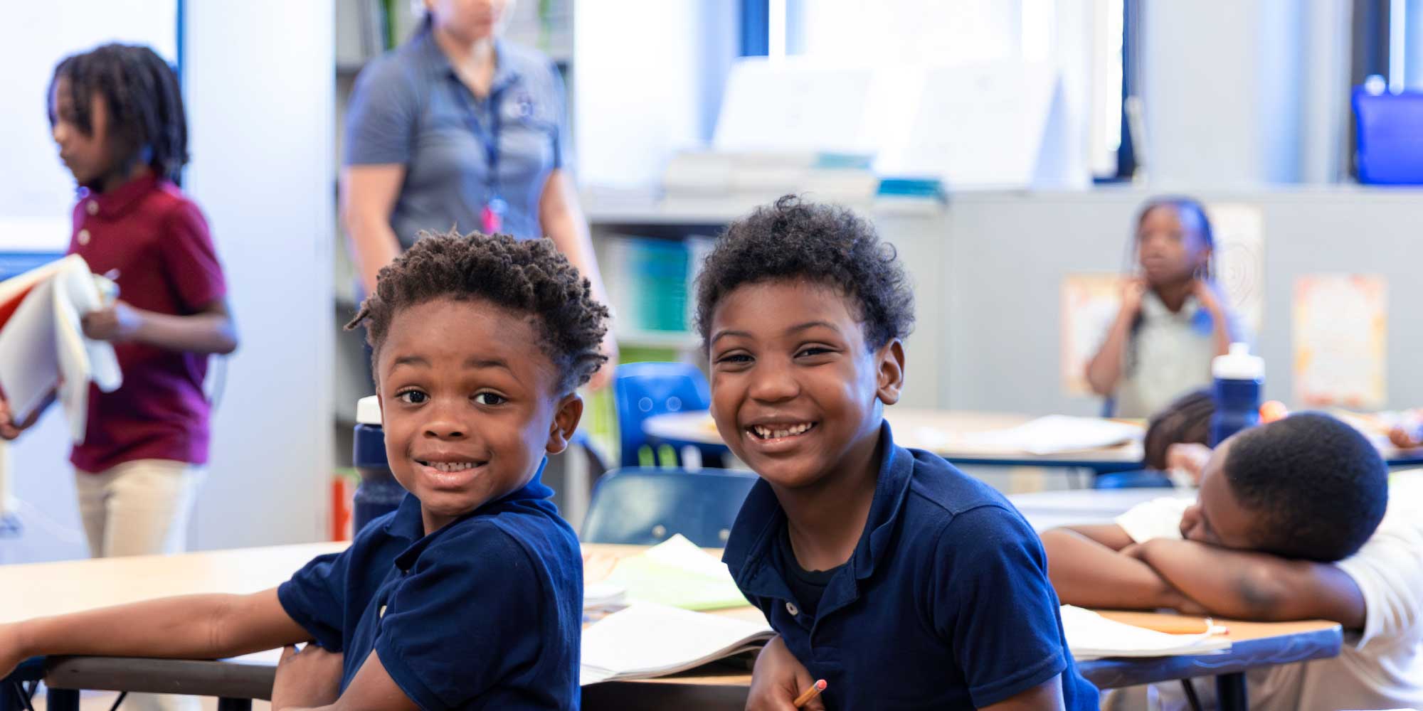 Two young students smiling at their desks while a teacher works with the class behind them.